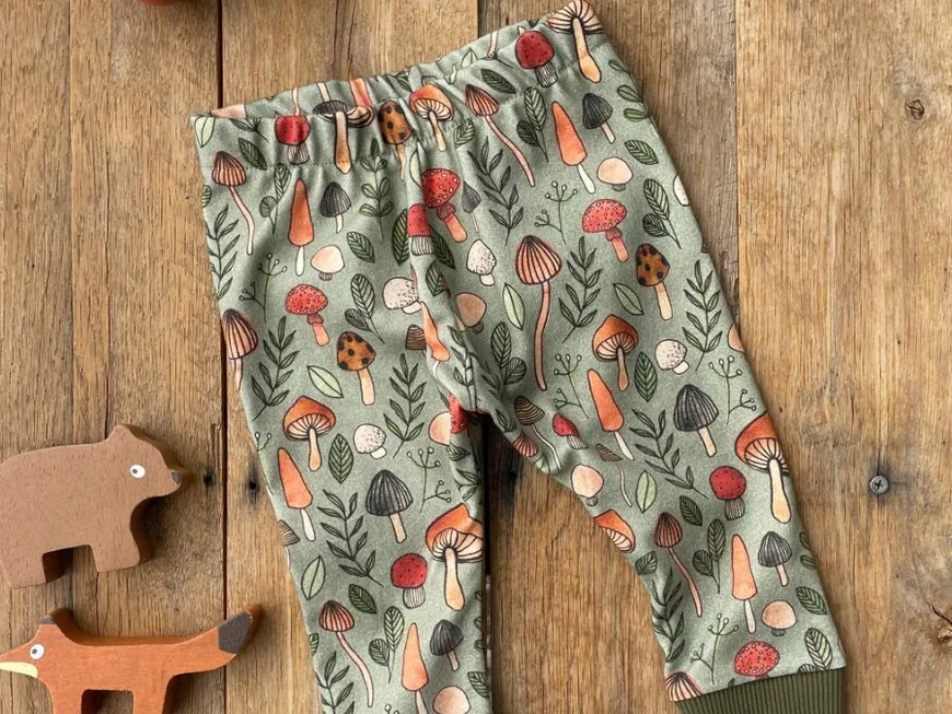 Baby pants in a green mushroom print laid on wood next to wooden animal toys
