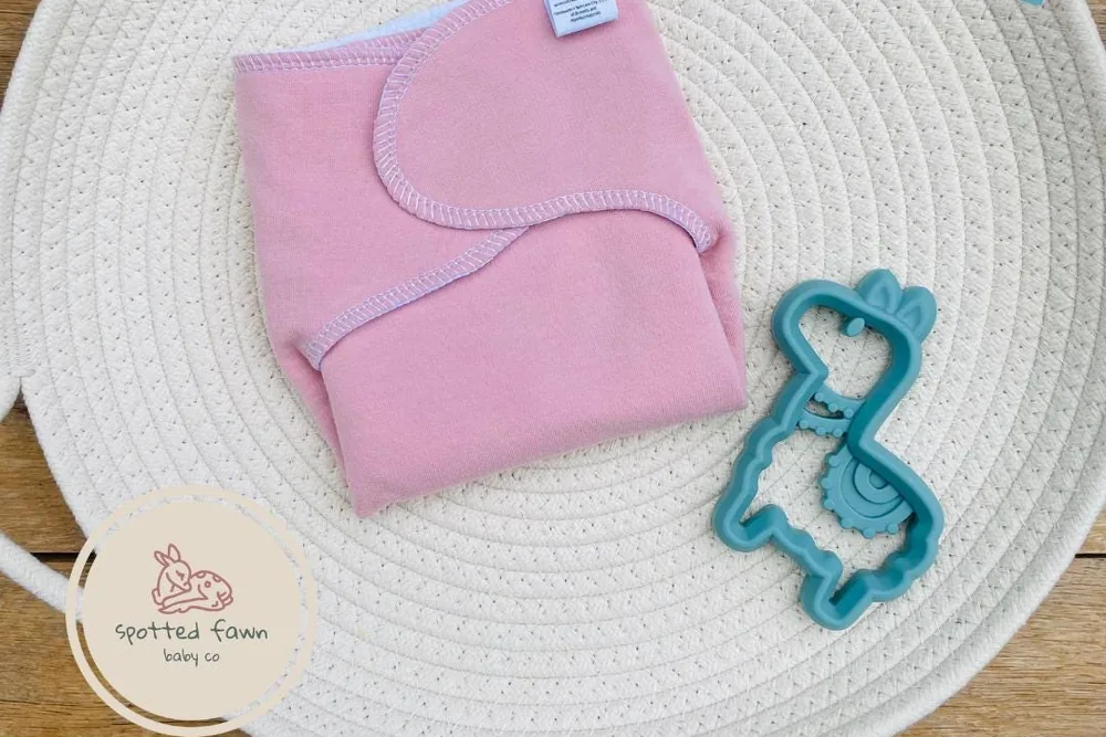 A pink cotton stretchy preflat folded in a woven rope basket, next to a teal toy
