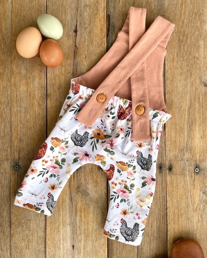 Crossed-back baby overalls in a floral chicken print with peach straps and wooden buttons, next to three fresh eggs